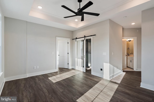 unfurnished bedroom featuring a barn door, recessed lighting, baseboards, wood finished floors, and a tray ceiling