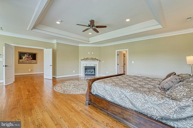 bedroom featuring a fireplace with flush hearth, a raised ceiling, light wood-style flooring, and crown molding
