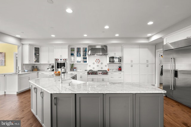 kitchen featuring stainless steel appliances, a large island with sink, decorative backsplash, gray cabinetry, and wall chimney range hood