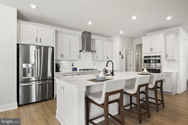 kitchen with wall chimney range hood, tasteful backsplash, a kitchen island with sink, stainless steel appliances, and light countertops