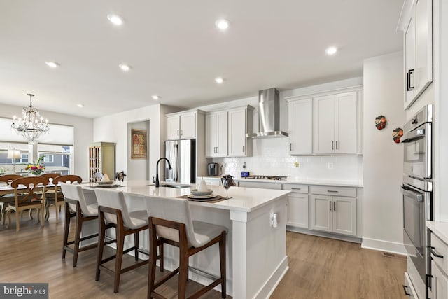 kitchen with wall chimney exhaust hood, stainless steel appliances, decorative backsplash, light countertops, and an island with sink