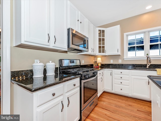 kitchen with appliances with stainless steel finishes, a sink, white cabinets, light wood-style floors, and dark stone counters