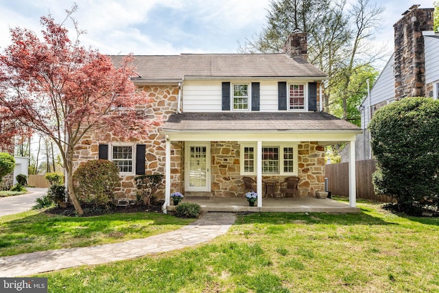 traditional-style house featuring covered porch, a chimney, fence, a front lawn, and stone siding
