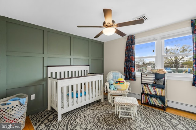 bedroom with visible vents, wood finished floors, a crib, a baseboard heating unit, and a decorative wall