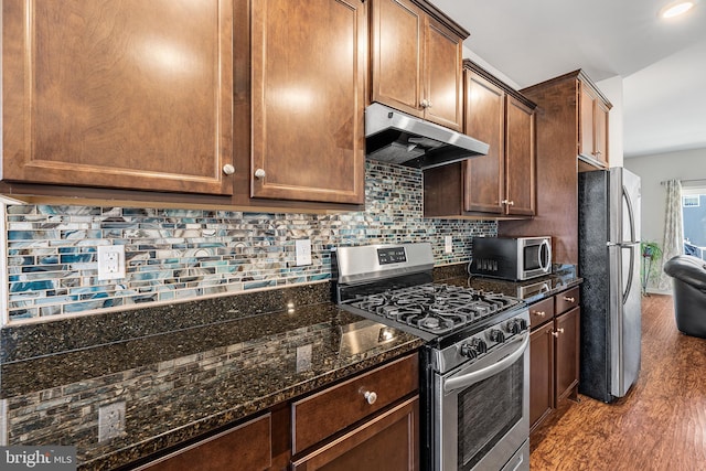 kitchen featuring appliances with stainless steel finishes, dark stone countertops, backsplash, wood finished floors, and under cabinet range hood