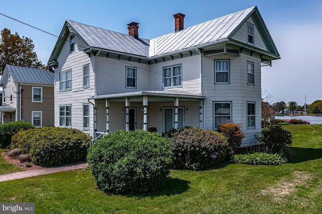 view of front facade featuring a chimney, metal roof, covered porch, a front yard, and a standing seam roof