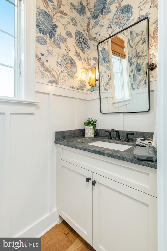 bathroom featuring wallpapered walls, wood finished floors, a wainscoted wall, and vanity