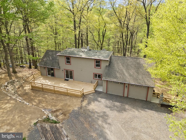 view of front of house with a deck, roof with shingles, an attached garage, driveway, and a forest view