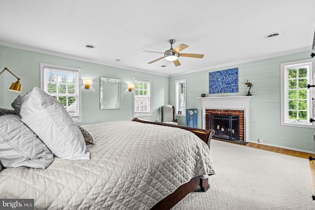 bedroom with wood finished floors, multiple windows, a brick fireplace, ornamental molding, and a ceiling fan