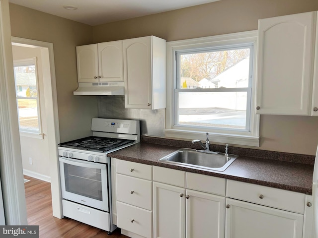 kitchen with dark countertops, a sink, white range with gas stovetop, white cabinetry, and under cabinet range hood