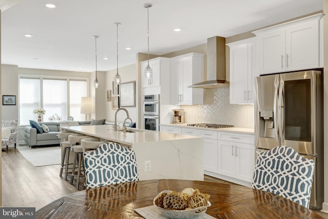 kitchen with wall chimney range hood, appliances with stainless steel finishes, a sink, a center island with sink, and open floor plan