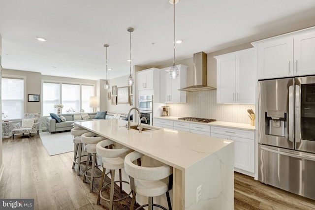 kitchen with stainless steel appliances, wall chimney range hood, wood finished floors, a breakfast bar area, and an island with sink