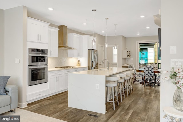 kitchen featuring appliances with stainless steel finishes, wall chimney range hood, a sink, wood finished floors, and an island with sink