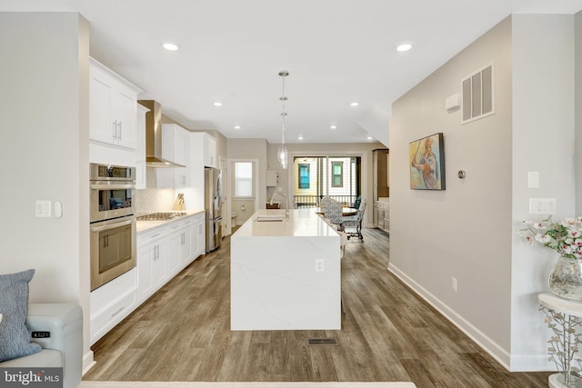 kitchen featuring wall chimney exhaust hood, a sink, appliances with stainless steel finishes, a kitchen island with sink, and baseboards
