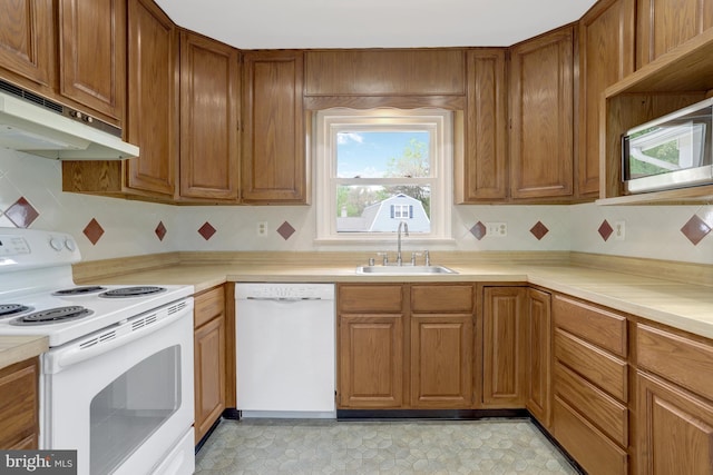 kitchen featuring brown cabinetry, a sink, light countertops, under cabinet range hood, and white appliances