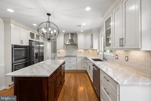 kitchen with wall chimney range hood, appliances with stainless steel finishes, a sink, a notable chandelier, and white cabinetry