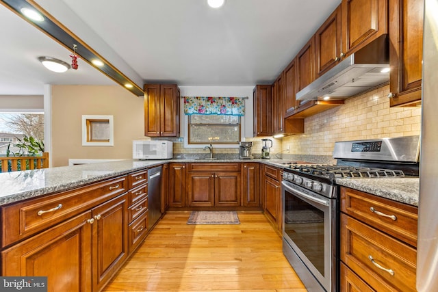 kitchen featuring light stone counters, stainless steel appliances, light wood finished floors, backsplash, and under cabinet range hood