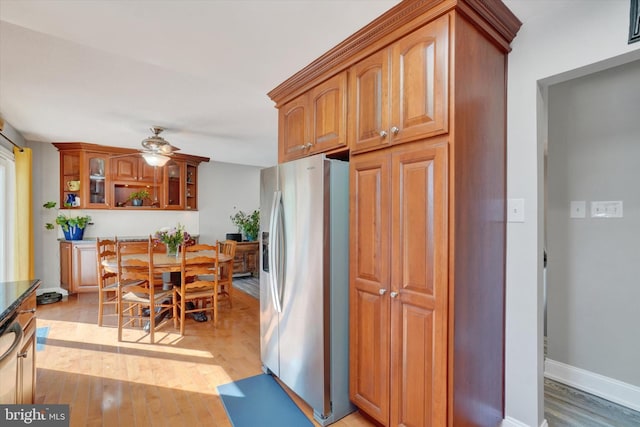 kitchen with glass insert cabinets, brown cabinetry, a ceiling fan, stainless steel refrigerator with ice dispenser, and light wood-style floors