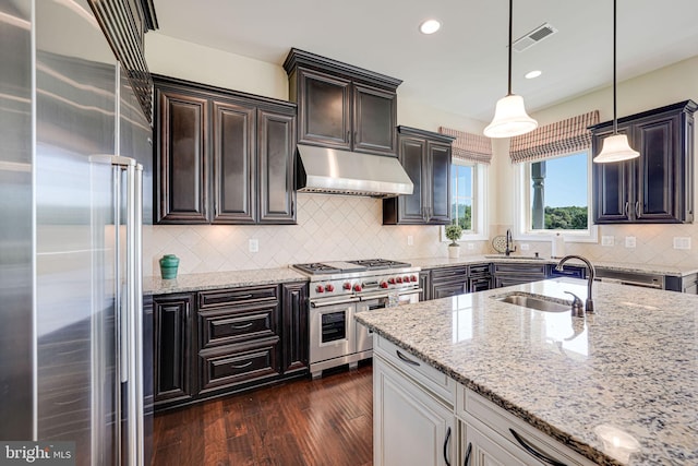 kitchen with under cabinet range hood, premium appliances, light stone counters, a sink, and tasteful backsplash