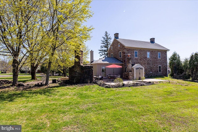 back of property featuring a chimney, an outdoor structure, a storage unit, a patio, and a lawn
