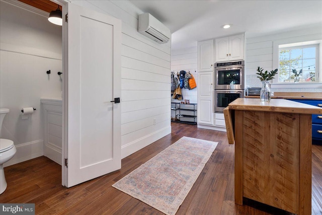 kitchen featuring stainless steel double oven, a wall mounted AC, dark wood-type flooring, white cabinetry, and baseboards