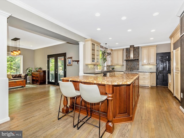kitchen featuring wall chimney exhaust hood, cream cabinets, light wood-style floors, a kitchen bar, and glass insert cabinets