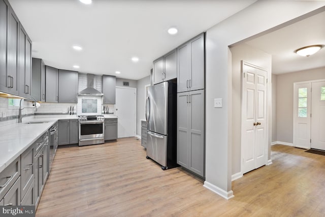 kitchen featuring appliances with stainless steel finishes, wall chimney exhaust hood, gray cabinetry, a sink, and light wood finished floors