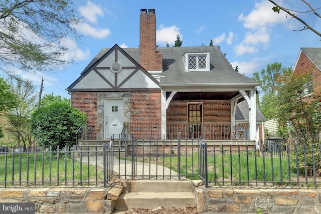 tudor house featuring a chimney, brick siding, a porch, and a fenced front yard