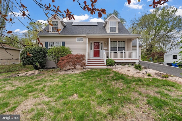 cape cod-style house featuring a shingled roof, a porch, a front lawn, and roof mounted solar panels
