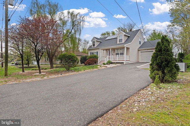 cape cod house featuring driveway, roof mounted solar panels, a shingled roof, covered porch, and a garage
