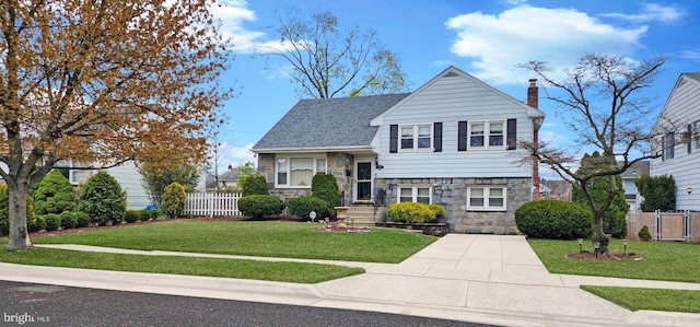 split level home featuring fence, a chimney, stone siding, and a front lawn