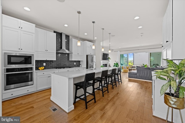 kitchen featuring stainless steel appliances, wall chimney range hood, open floor plan, light countertops, and light wood-style flooring