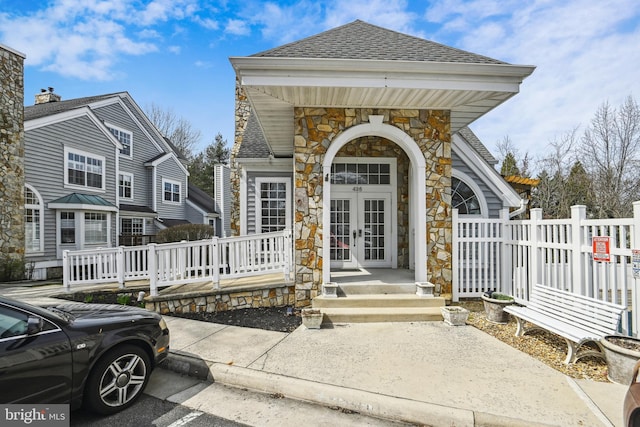 doorway to property with a shingled roof, covered porch, stone siding, french doors, and fence