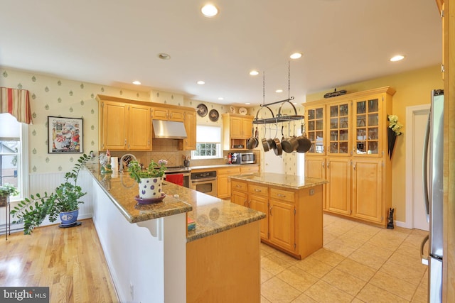kitchen featuring light stone countertops, under cabinet range hood, a peninsula, a kitchen island, and appliances with stainless steel finishes