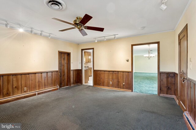 carpeted empty room featuring track lighting, wood walls, wainscoting, and ceiling fan with notable chandelier