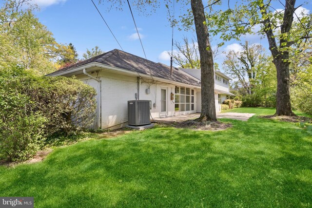 rear view of house with a shingled roof, central AC, brick siding, a patio area, and a lawn