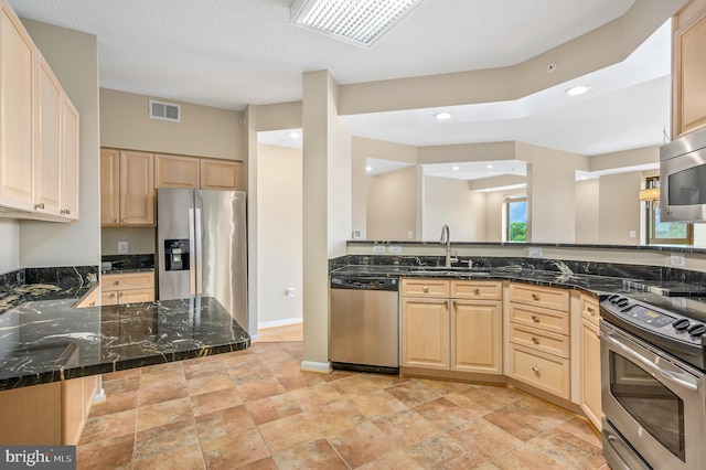 kitchen featuring appliances with stainless steel finishes, light brown cabinetry, dark stone counters, a sink, and recessed lighting