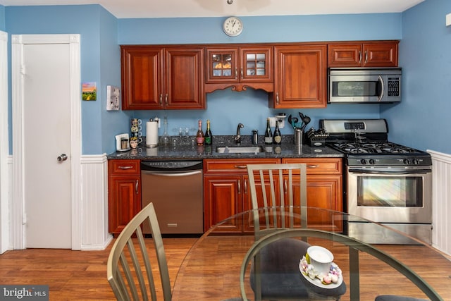 kitchen with a sink, dark stone countertops, light wood-type flooring, a wainscoted wall, and appliances with stainless steel finishes