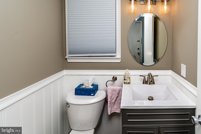 half bath with wainscoting, toilet, a decorative wall, and vanity