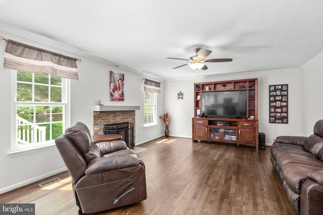 living room featuring dark wood-style flooring, ceiling fan, a stone fireplace, baseboards, and ornamental molding