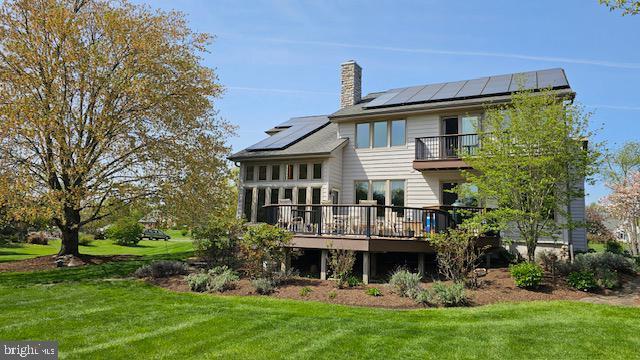 back of house with a chimney, a standing seam roof, roof mounted solar panels, a balcony, and a lawn