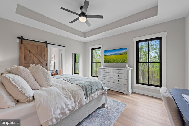 bedroom with baseboards, a barn door, a tray ceiling, a ceiling fan, and light wood-style flooring