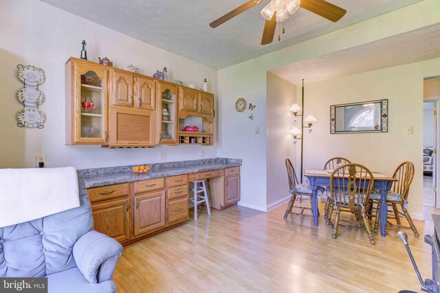 kitchen featuring ceiling fan, light wood finished floors, brown cabinetry, glass insert cabinets, and dark countertops