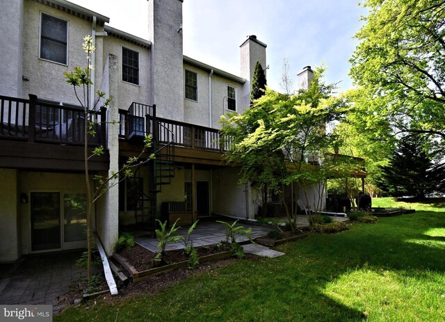 back of house featuring a chimney, a wooden deck, a yard, a patio, and stucco siding