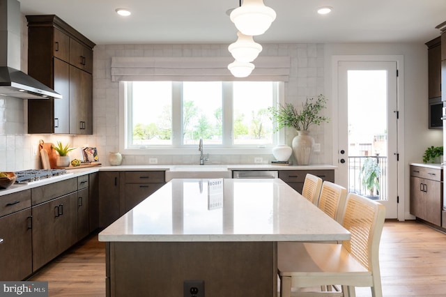 kitchen with a sink, light wood-style floors, dark brown cabinets, wall chimney exhaust hood, and backsplash