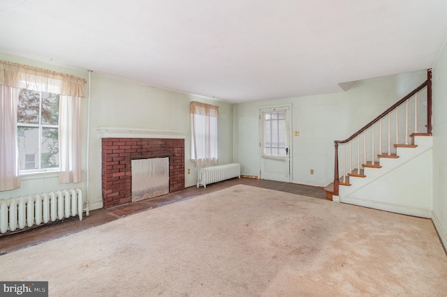unfurnished living room featuring a brick fireplace, carpet, stairs, and radiator