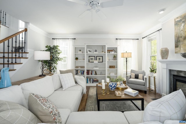 living area featuring a glass covered fireplace, stairs, a ceiling fan, wood finished floors, and ornamental molding