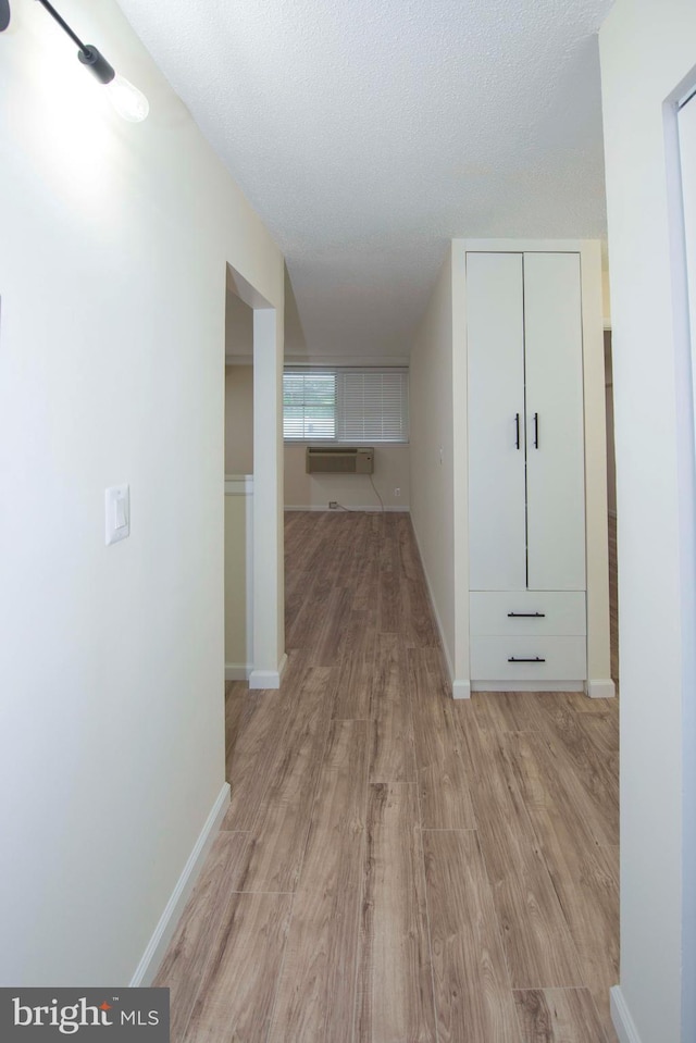 hallway featuring a textured ceiling, light wood-type flooring, a wall unit AC, and baseboards