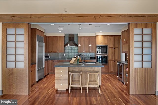 kitchen with dark countertops, wall chimney exhaust hood, brown cabinets, a sink, and appliances with stainless steel finishes