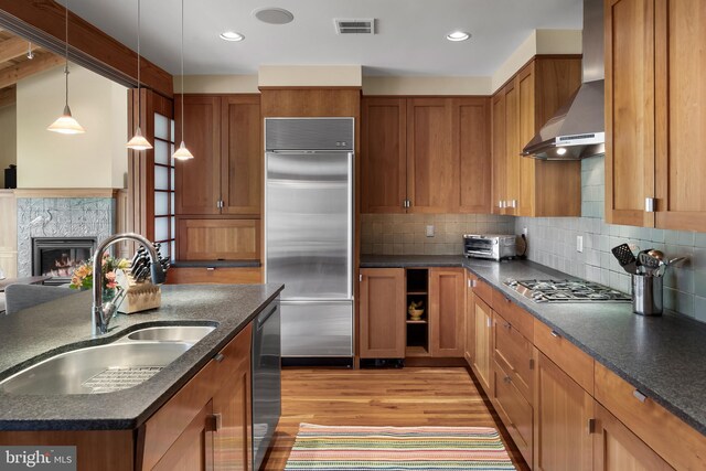 kitchen with brown cabinetry, appliances with stainless steel finishes, wall chimney exhaust hood, a sink, and light wood-style flooring
